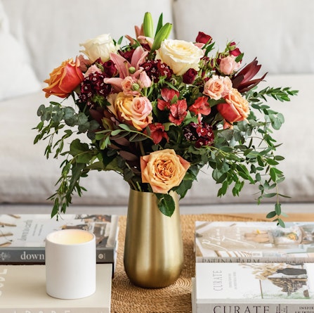 A vibrant floral arrangement featuring roses, daisies, and greenery in a stylish gold vase.