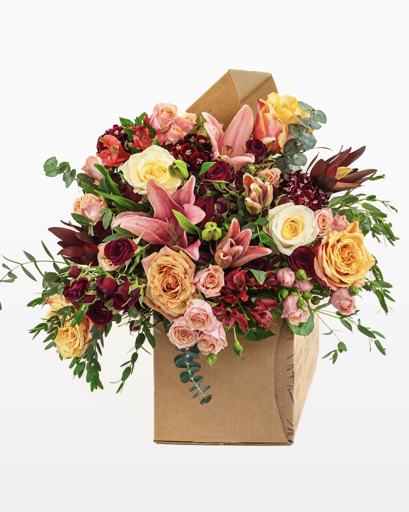 Vibrant bouquet of mixed flowers including roses, lilies, and green foliage, packaged in a craft paper cone, displayed against a white background.