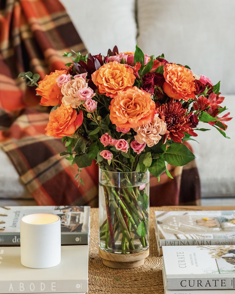 A vibrant bouquet of orange roses and dark red flowers in a clear glass vase, placed on a table with books and a lit candle, giving a cozy, homey atmosphere.
