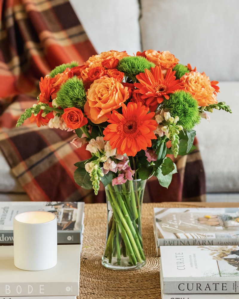Vibrant bouquet of orange roses and gerbera daisies with green accent flowers in a clear vase on a coffee table, with a candle and magazines nearby.