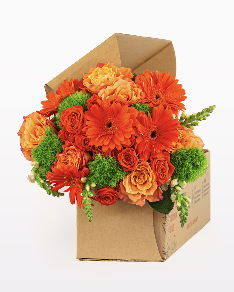 Vibrant bouquet of orange flowers, including gerberas and roses, with green accents, artistically arranged in a brown cardboard box against a white background.