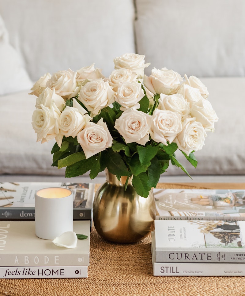 Bouquet of white roses in a golden vase on a coffee table with books and a lit candle, creating a cozy and elegant home interior setting.
