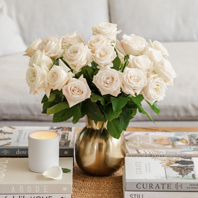 Bouquet of white roses in a golden vase on a coffee table with books and a lit candle, creating a cozy and elegant home interior setting.