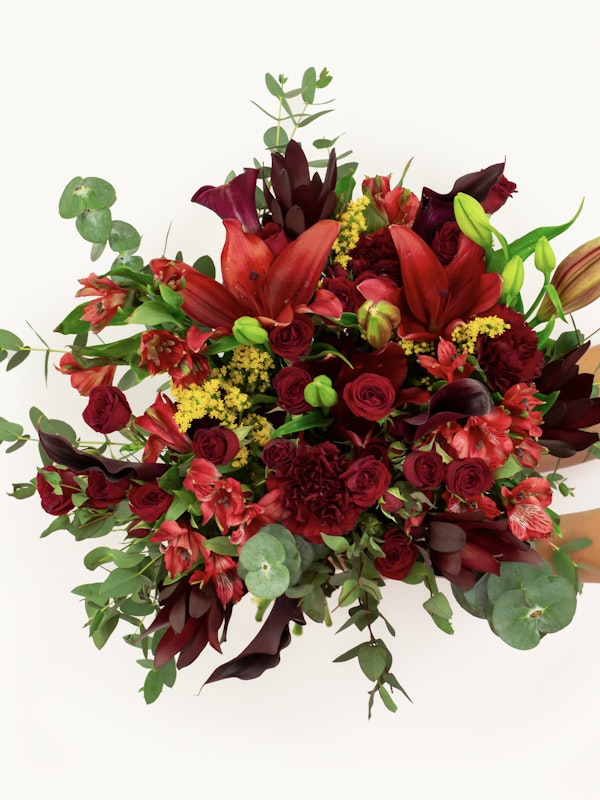 A vibrant bouquet with red lilies, deep red roses, and various greenery against a white background, held by a person with a subtle glimpse of hands.