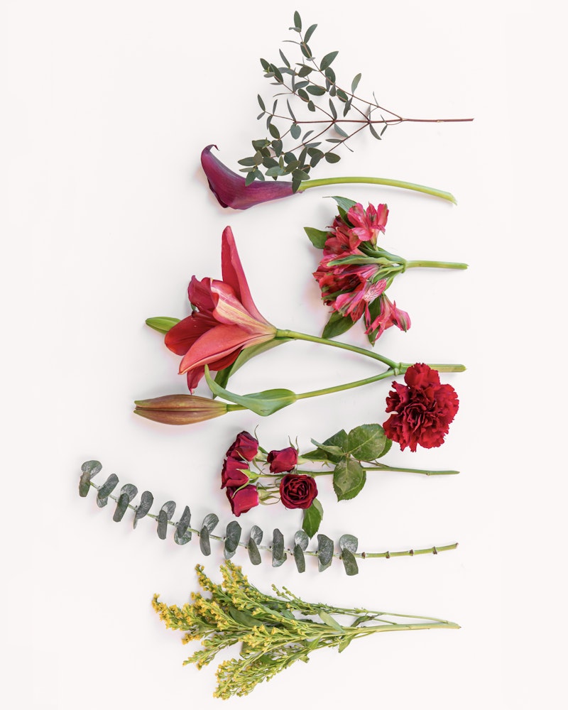 Assorted fresh flowers and greenery arranged in a vertical line on a white background, featuring red lilies, pink blossoms, carnations, and eucalyptus.