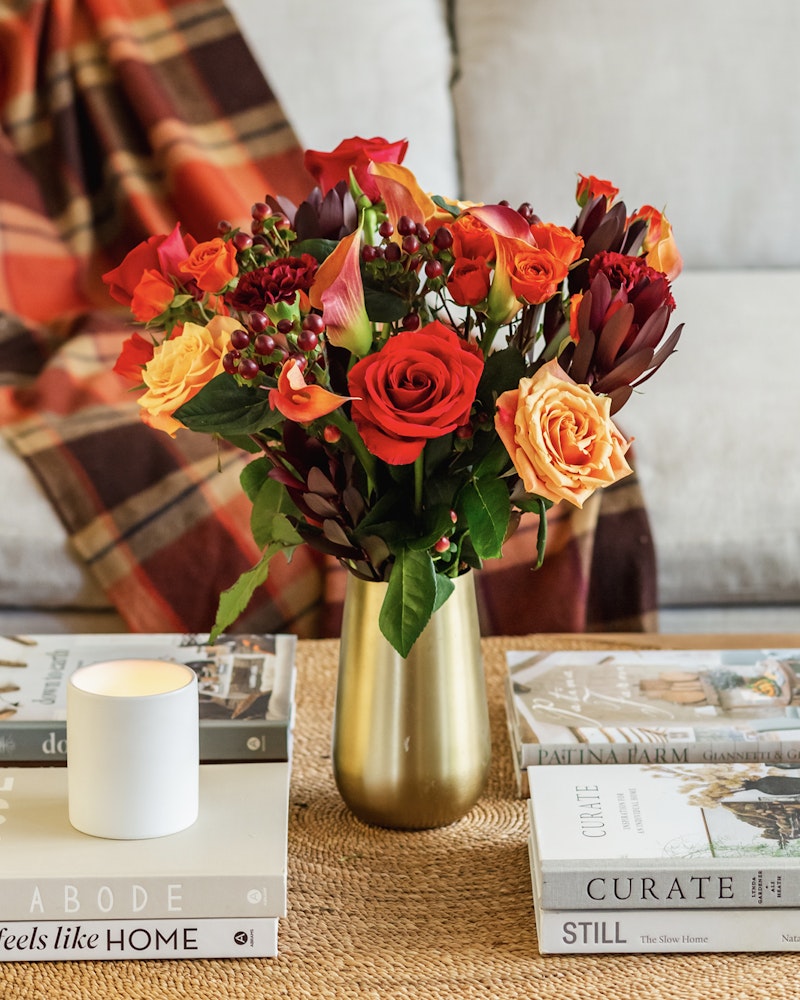 Vibrant bouquet of red flowers in a gold vase on a coffee table with books and a lit candle, creating a cozy living room atmosphere.