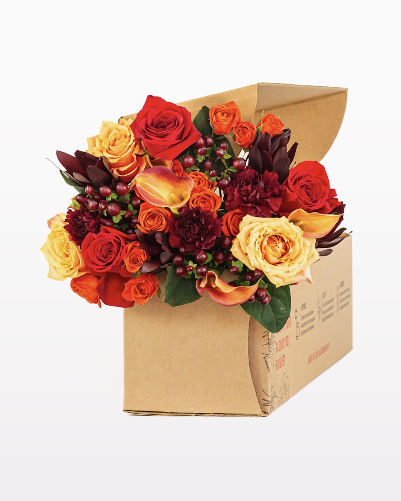 Vibrant bouquet of red and yellow roses, orange lilies, and purple accents in a cardboard flower box against a white background, showcasing a fresh floral delivery concept.