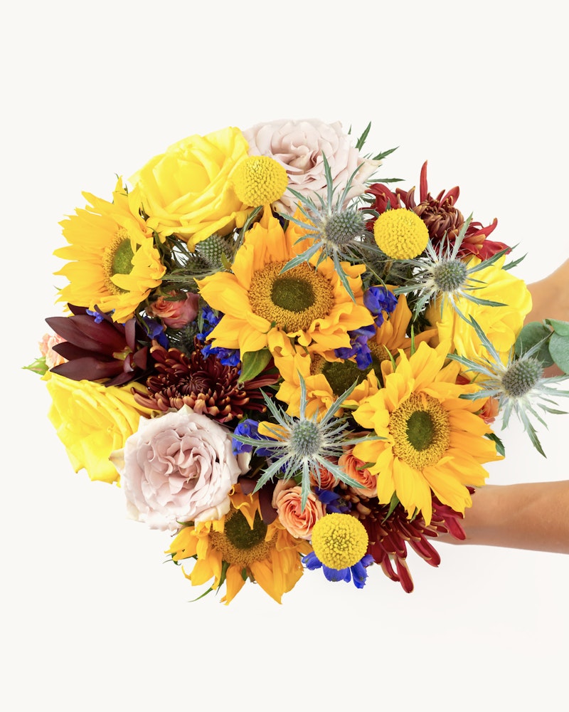 Vibrant bouquet of flowers including sunflowers, roses, and other colorful blooms held against a white background, showcasing a diverse and bright floral arrangement.