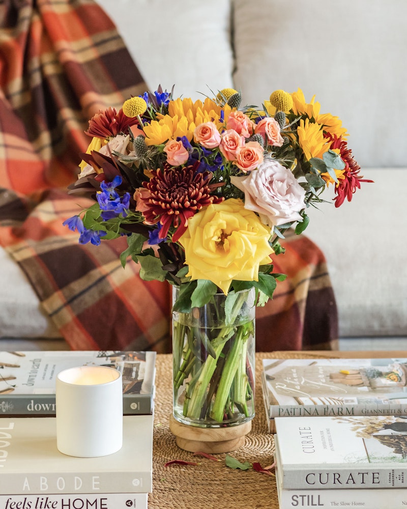 Vibrant bouquet of assorted flowers in a clear glass vase on a coffee table with books and a lit candle, complemented by a cozy plaid throw in the background.