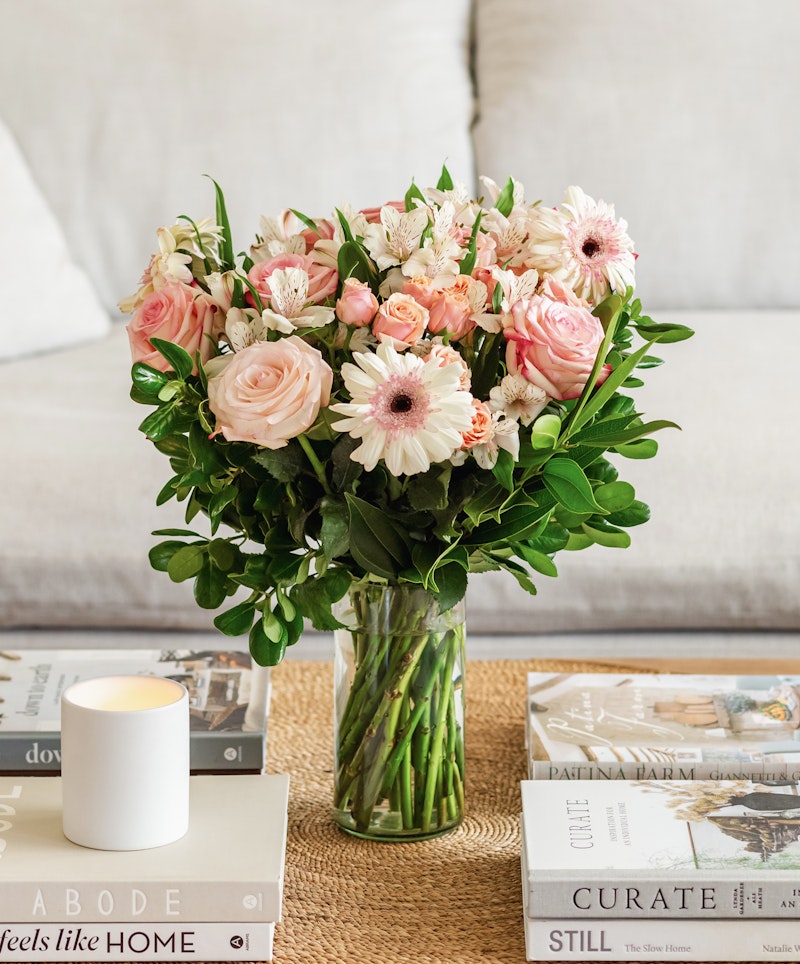 Beautiful bouquet of pink roses and white flowers in a glass vase on a coffee table with books and a lit candle, giving a cozy and inviting home atmosphere.