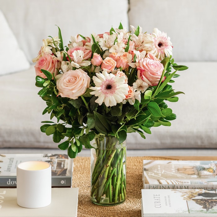 Beautiful bouquet of pink roses and white flowers in a glass vase on a coffee table with books and a lit candle, giving a cozy and inviting home atmosphere.