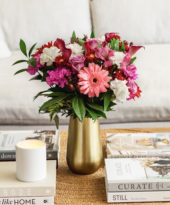 Vibrant bouquet of mixed flowers in a gold vase on a coffee table with books and a white candle, in a cozy living room setting.