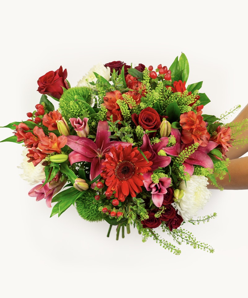 Vibrant bouquet of flowers with red, pink, and white blooms including roses and lilies, held in hands against a white background, showcasing a variety of textures and greenery.