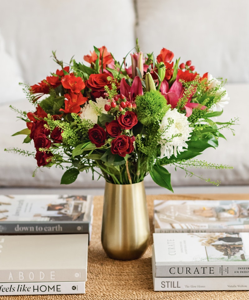 Vibrant bouquet of red roses, lilies, and assorted greenery in a gold vase on a table with stacked decor books in a cozy living room setting.