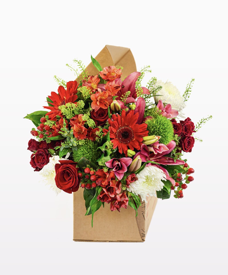 Vibrant bouquet of flowers with red roses, gerberas, and mixed blooms, with greenery in a brown paper wrapper against a white background.