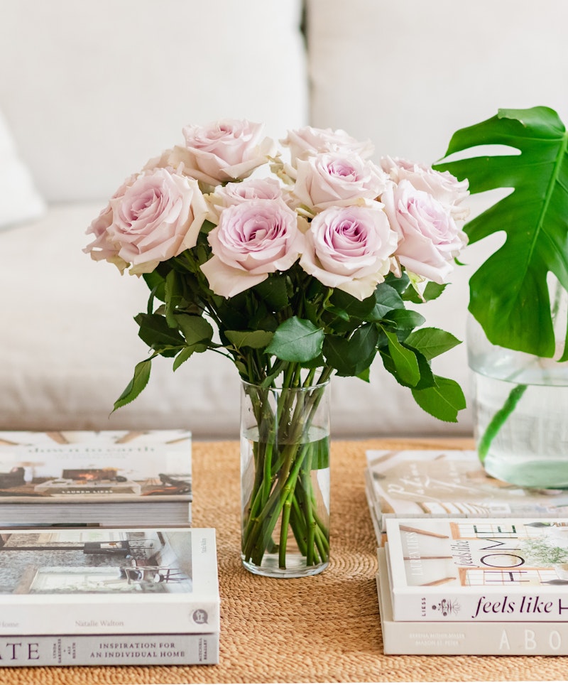 A bouquet of delicate pink roses in a clear vase, placed on a woven mat surrounded by an assortment of hardcover books on a cozy living room table.