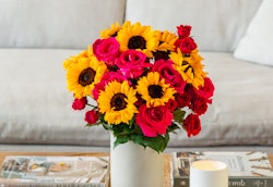 Vibrant bouquet of yellow sunflowers and red roses in a white vase on a wooden coffee table with a cozy grey sofa and magazines in the background.