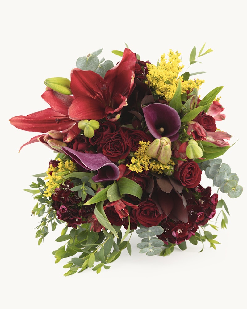Elegant bouquet featuring deep red lilies, burgundy roses, purple calla lilies, with sprigs of yellow solidago and green eucalyptus leaves on a white background.