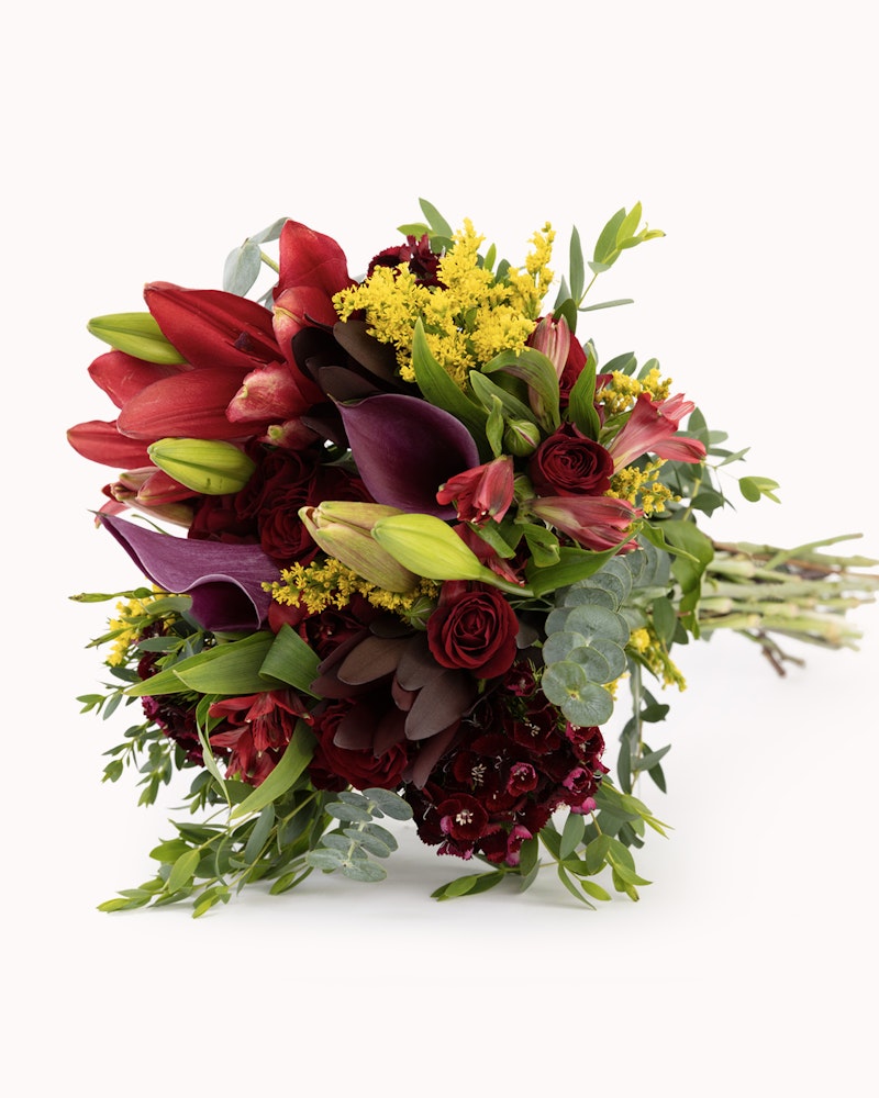 Vibrant bouquet of red and burgundy flowers, including lilies and roses, with yellow filler blooms and green foliage against a white background.