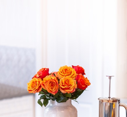 A vibrant bouquet of orange roses in a white vase on a table, with a blurred background featuring a cozy room with a grey sofa and a modern silver teapot.