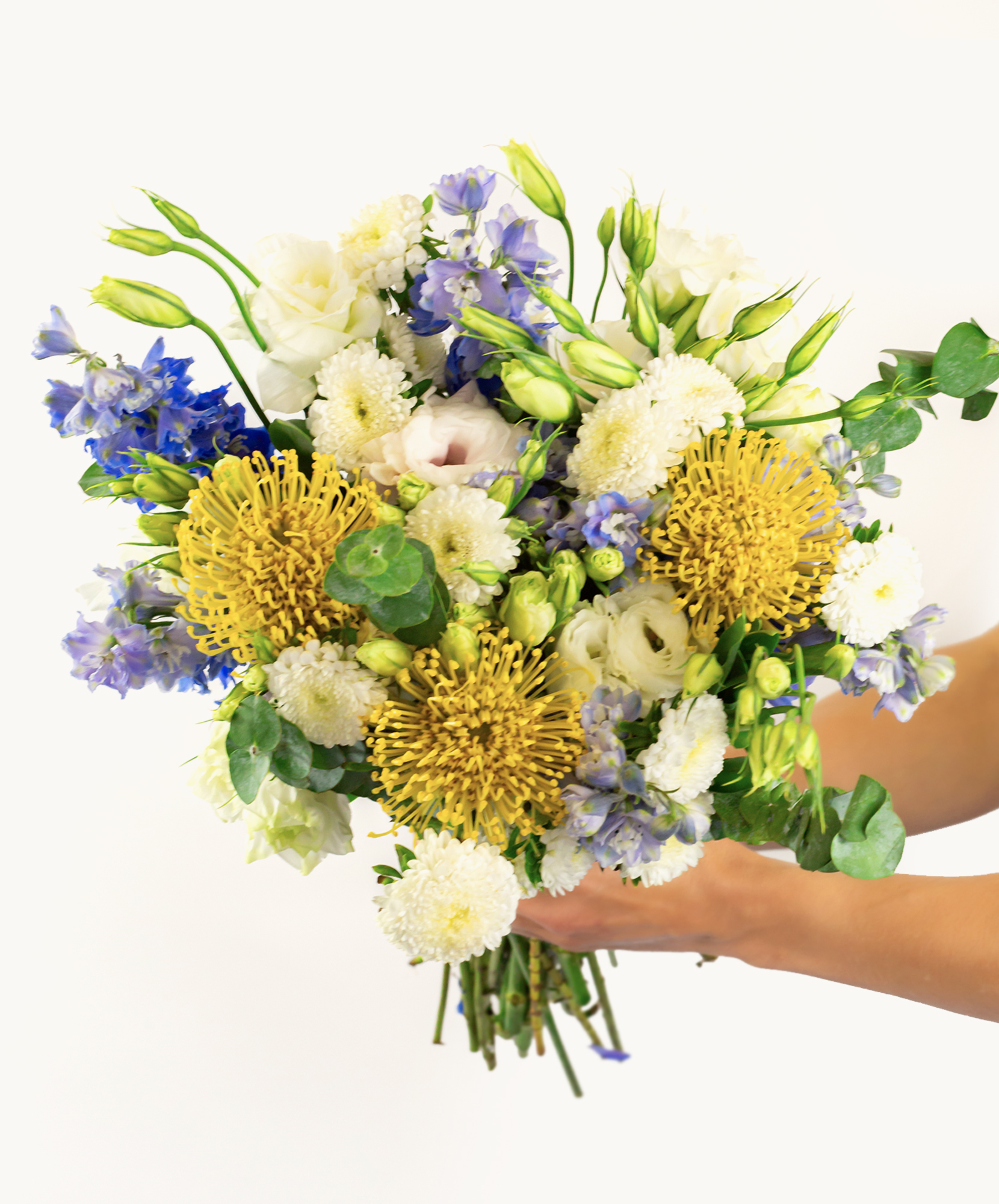 A vibrant bouquet featuring yellow proteas, blue delphiniums, and white blooms, elegantly held.