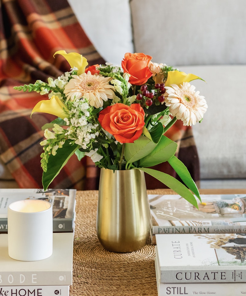 A vibrant bouquet of yellow lilies, orange roses, and white daisies in a golden vase on a table with books and a lit candle, creating a cozy home setting.