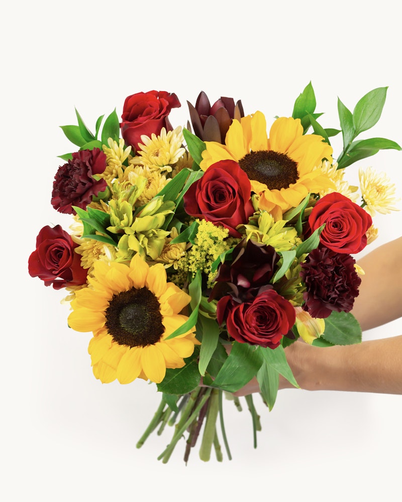 A vibrant bouquet of flowers presented against a white background, featuring red roses, yellow sunflowers, and an assortment of complementary blooms held by a person.