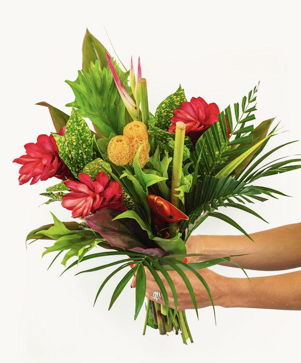 Vibrant tropical bouquet held by a person's arm against a white background, featuring red ginger flowers, yellow sponge, and lush green leaves.