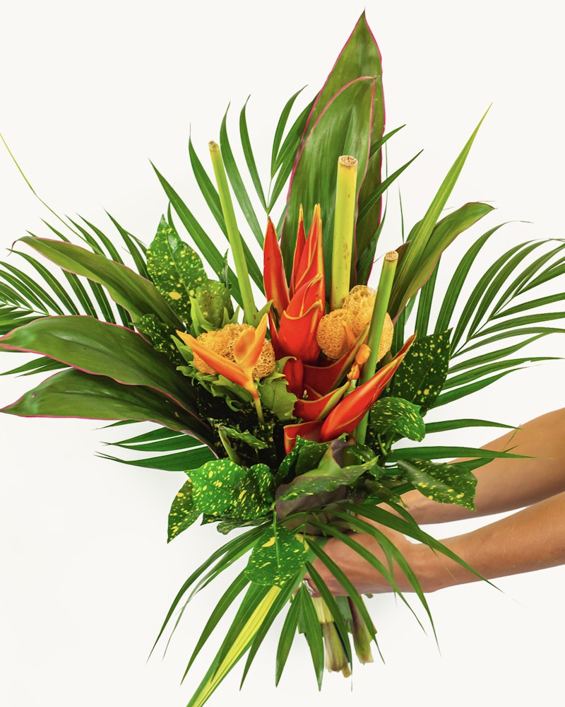Vibrant tropical bouquet with oranges and greens, featuring heliconia, anthurium, and lush foliage against a clean white background.