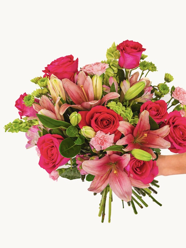 A person holds a vibrant bouquet of flowers featuring pink lilies, deep red roses, and various green leaves and stems, presented against a white background.