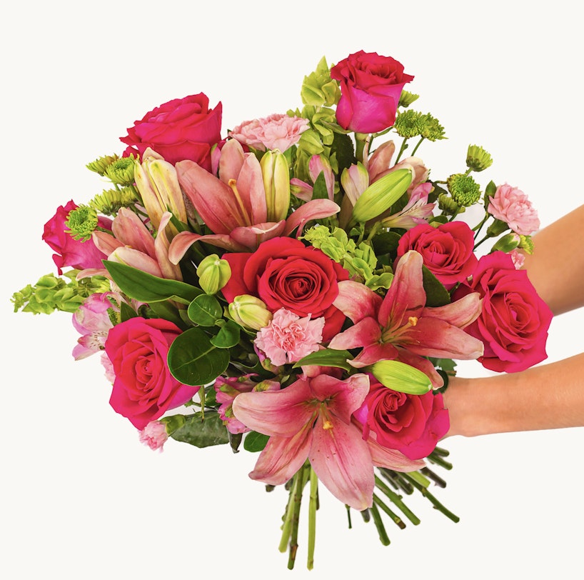 A person holds a vibrant bouquet of flowers featuring pink lilies, deep red roses, and various green leaves and stems, presented against a white background.