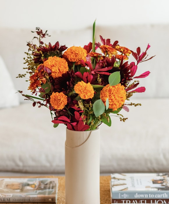 Vibrant bouquet of orange and red flowers arranged in a white vase on a coffee table with books and magazines, conveying a cozy, autumnal atmosphere.