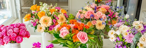 Panoramic view of a vibrant collection of assorted flowers including roses, daisies, and lilies in full bloom, displayed on a bright windowsill.