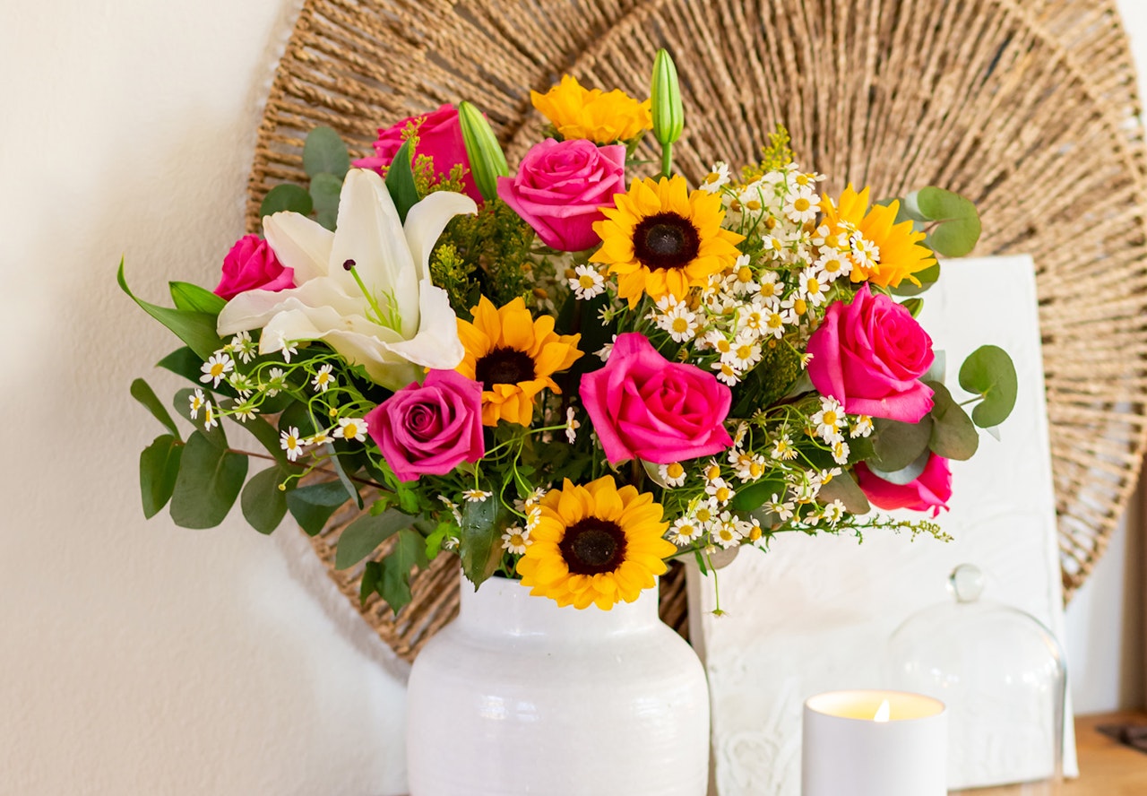 Beautiful bouquet of flowers including pink roses, white lilies, and sunflowers in a white vase, with a lit candle on a table against a textured wall.