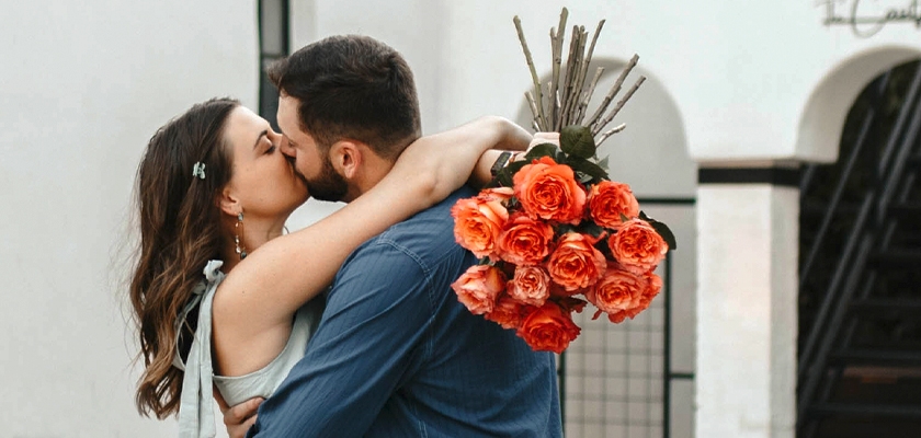 A couple sharing a romantic kiss, holding a vibrant bouquet of orange roses.