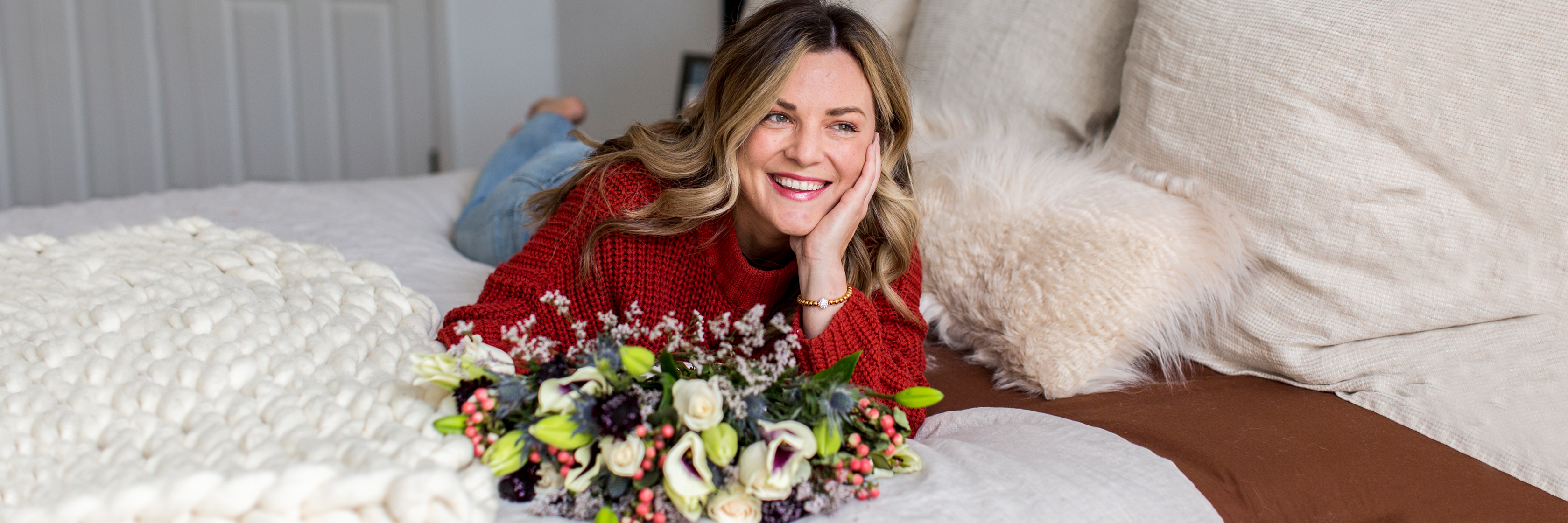 A cheerful woman in a cozy sweater smiles with a vibrant flower bouquet on her bed.