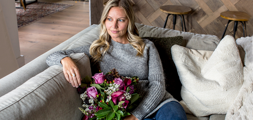 A woman sitting on a cozy sofa, holding a vibrant bouquet of pink flowers and greenery.
