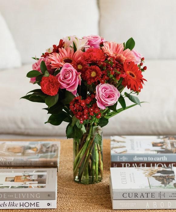 A vibrant bouquet of pink roses, gerberas, and red foliage arranged in a clear vase on a table with stylish home decor books.