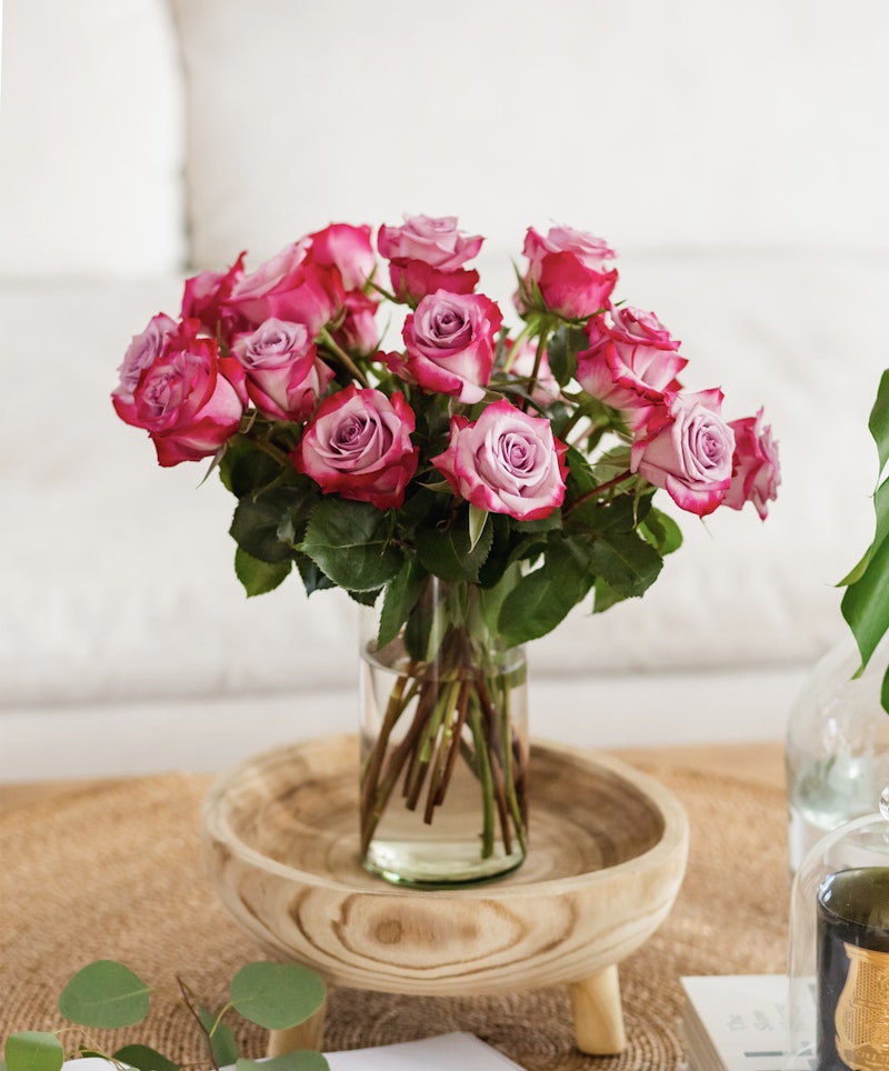 A bouquet of vibrant pink roses arranged in a clear glass vase on a wooden tray, with a cozy white cushioned background enhancing the warm ambiance.