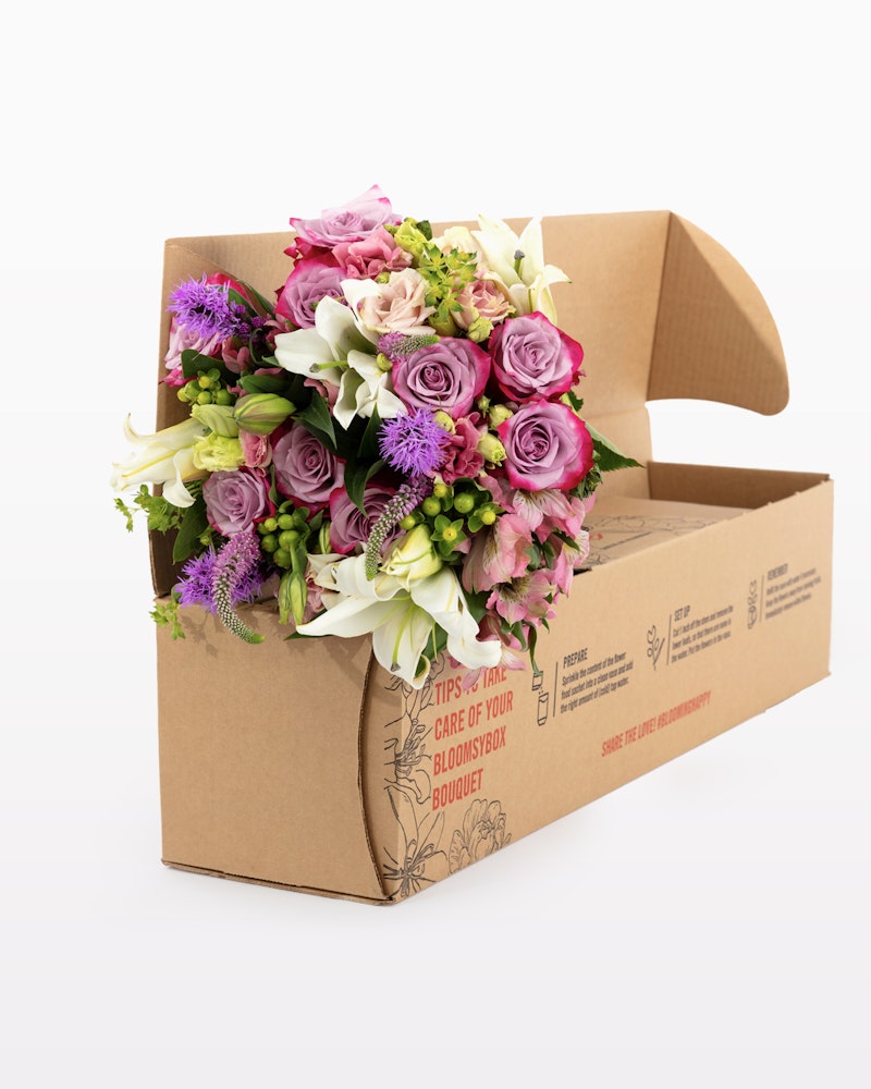 Vibrant bouquet of mixed flowers including roses and lilies in a cardboard box with care instructions, isolated on a white background, ready for delivery.