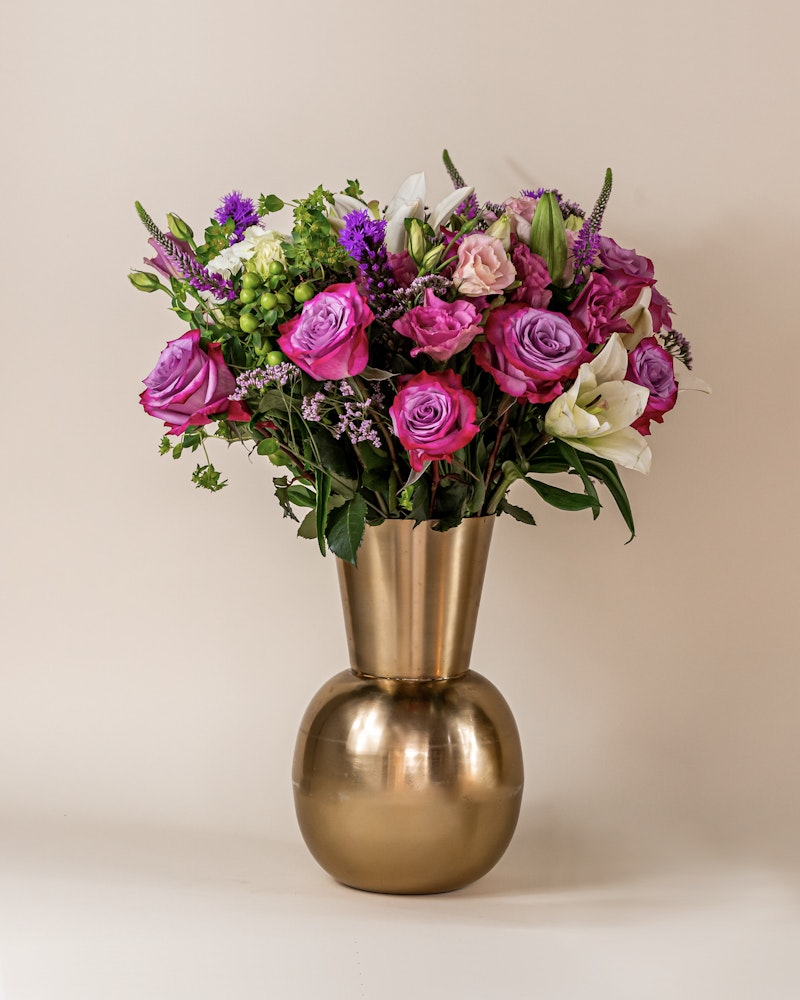 Vibrant bouquet of assorted flowers including purple roses and white lilies in a golden vase set against a neutral beige background, perfect for gifting or home decor.