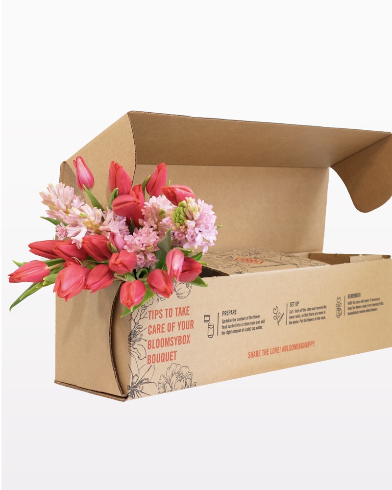 Bright pink and white flowers bloom from an open cardboard BloomsyBox, showcasing a floral bouquet ready for delivery with care instructions printed on the side.