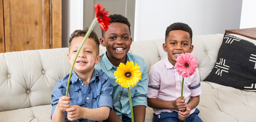 Three cheerful boys sitting on a couch, each holding colorful flowers, posing happily.