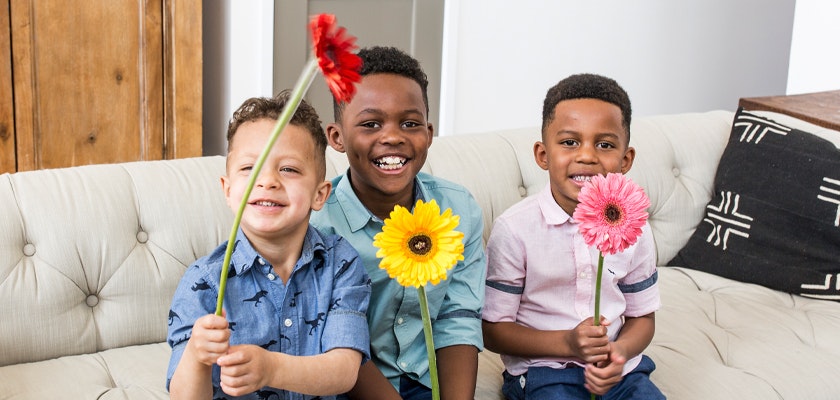 Three cheerful boys sitting on a couch, each holding colorful flowers, posing happily.