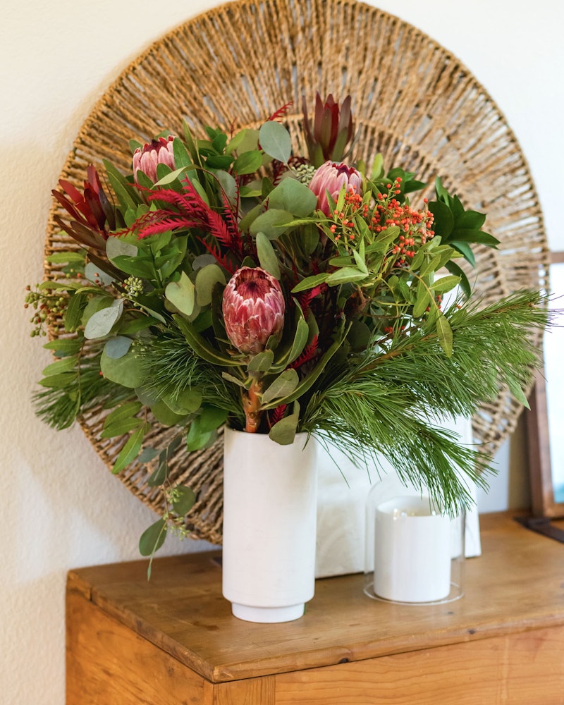 Vibrant bouquet of red protea flowers and green foliage in a white vase on a wooden table, with a circular woven wall decor in the background.
