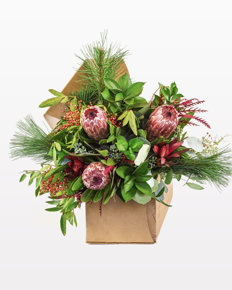 A vibrant bouquet featuring pink protea flowers, pine greenery, and mixed foliage presented in a crafted brown paper wrapping against a white background.