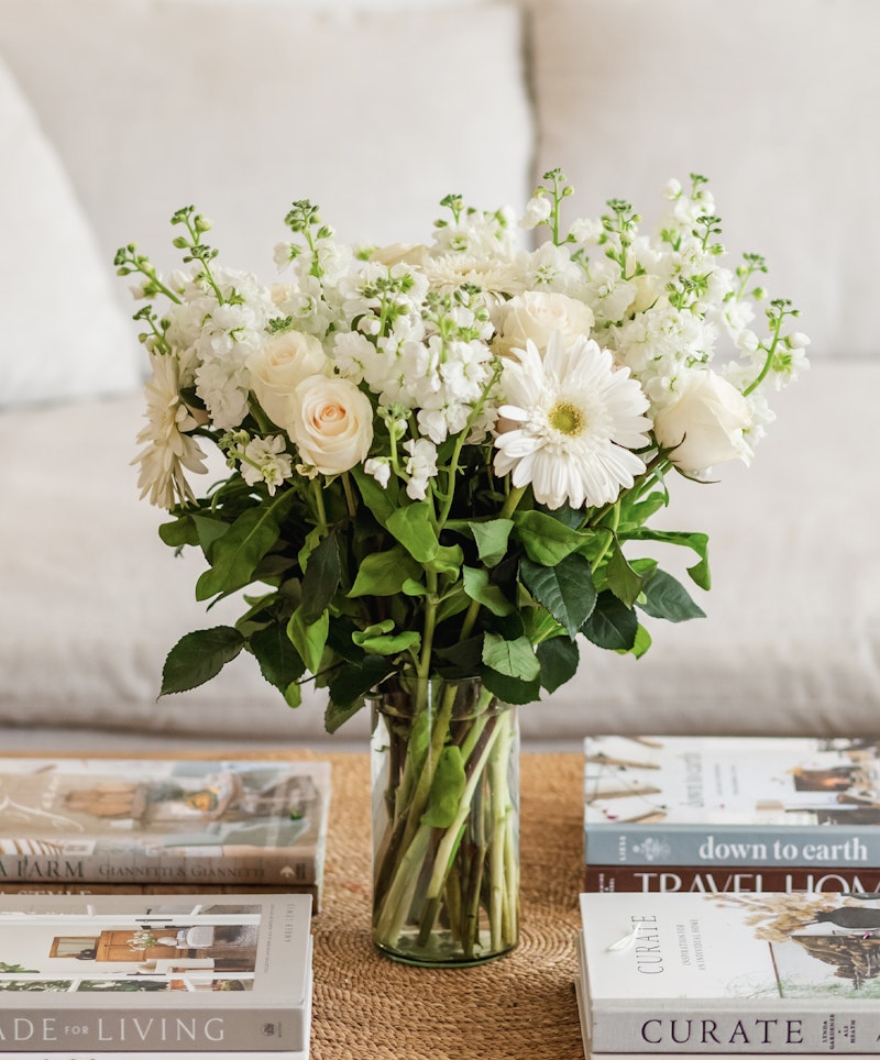 Elegant bouquet of white flowers, including roses and daisies, arranged in a clear glass vase on a table with stylish interior design books.