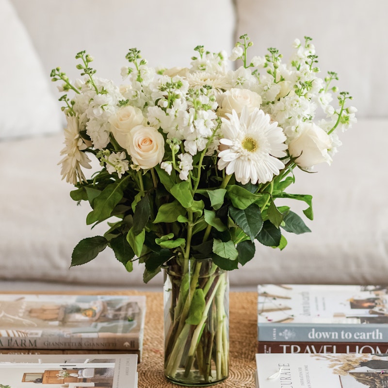 Elegant bouquet of white flowers, including roses and daisies, arranged in a clear glass vase on a table with stylish interior design books.