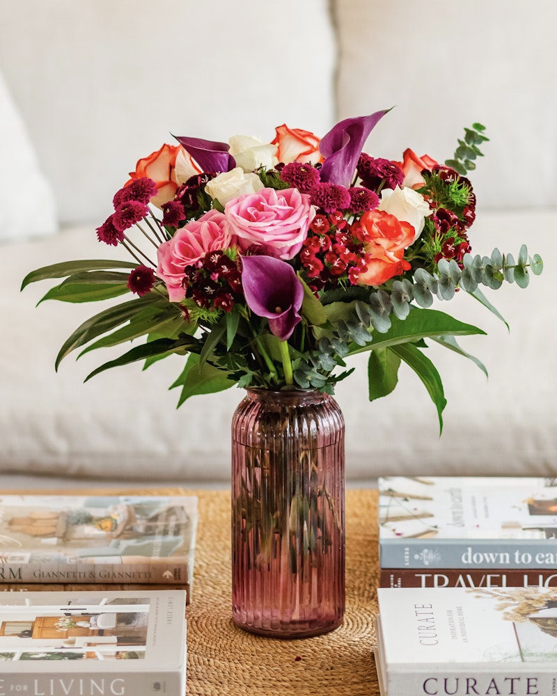 A vibrant bouquet of flowers with roses, lilies, and foliage in a pink vase on a coffee table with assorted books on interior design and travel.