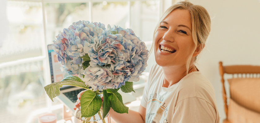 A smiling woman holds a vibrant bouquet of blue hydrangeas in a cozy indoor setting.